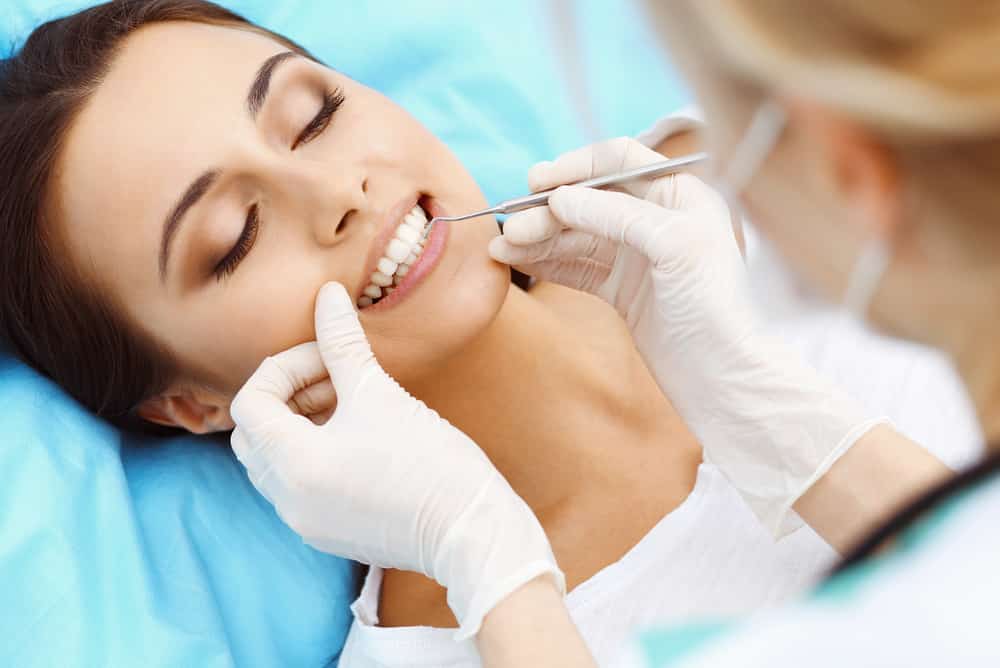 Young female patient visiting dentist office.Beautiful woman with healthy straight white teeth sitting at dental chair with open mouth during oral checkup while doctor working at teeth. Dental clinic, stomatology concept